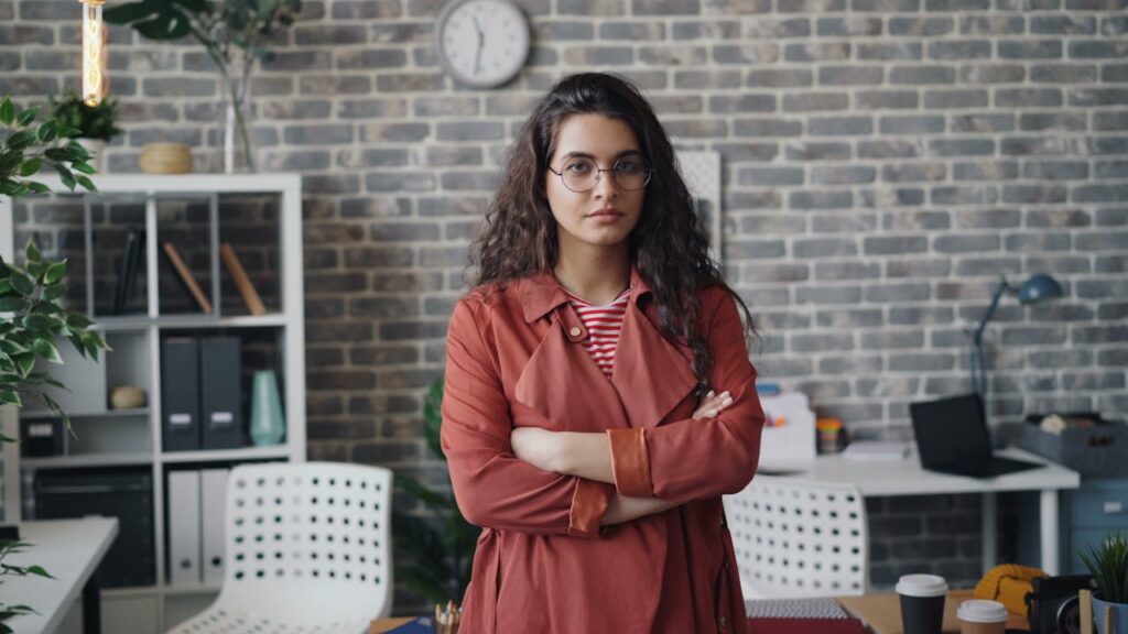 pexels-photo-23496712 A confident woman with crossed arms in a contemporary office setting, wearing eyeglasses and a red coat.