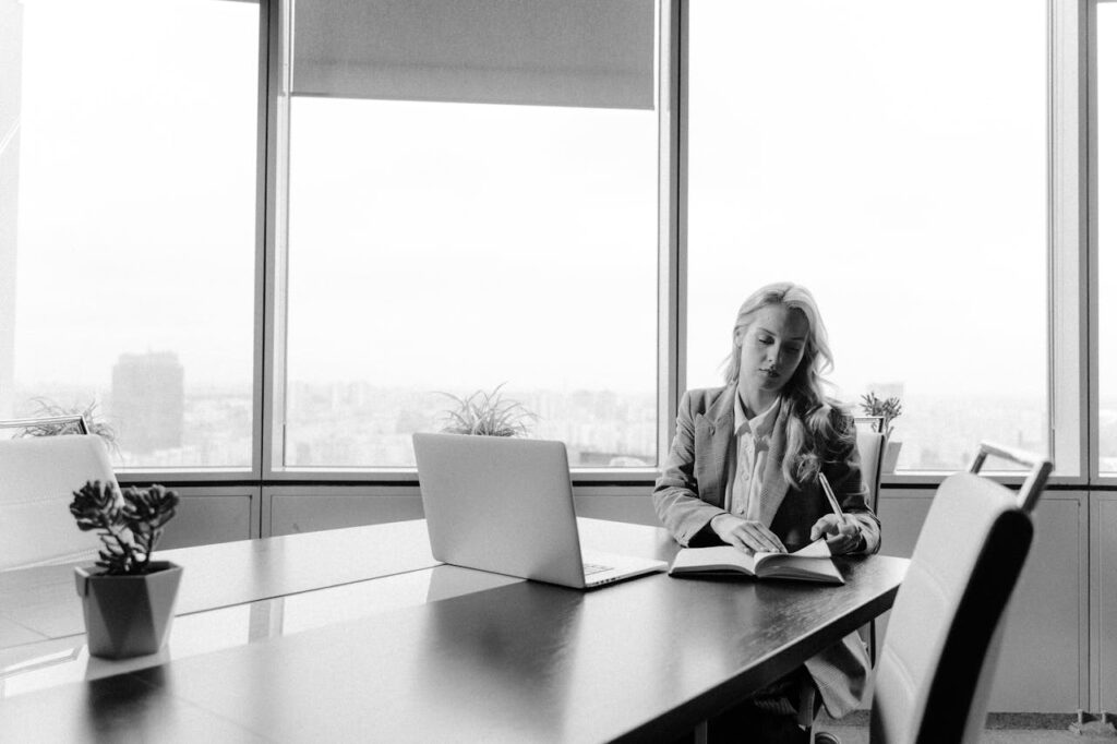 pexels-photo-5717562 Businesswoman working at a desk in a bright office with city view, in black and white.