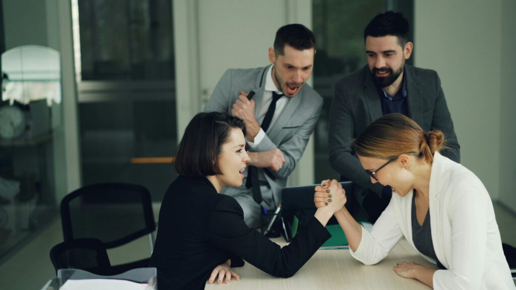 pexels-photo-36714224-36714224 Two female colleagues arm wrestling in an office setting with male coworkers cheering.