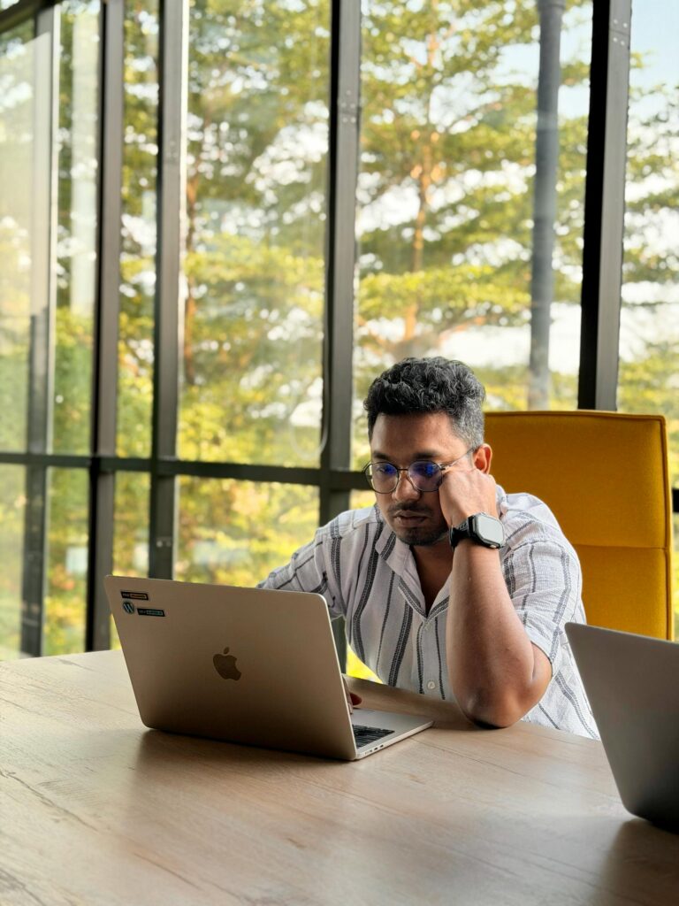 pexels-photo-37573719-37573719 Man in striped shirt working on a laptop at a contemporary office with large windows.