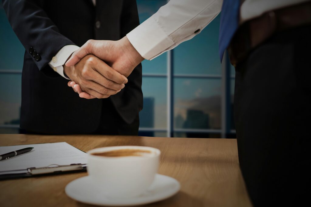 pexels-photo-4175026-4175026 Two professionals shaking hands across a desk with cityscape in the background.