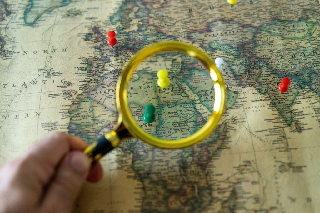 pexels-photo-12932264-12932264 Close-up of a hand holding a magnifying glass over a world map with colorful pins indicating travel destinations.